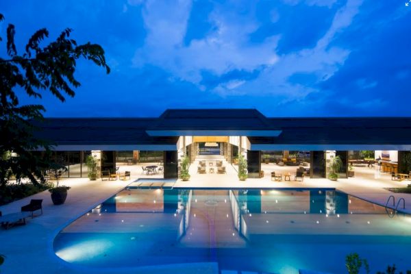 A luxurious poolside view of a modern building under a twilight sky, reflecting lights and surrounded by outdoor seating and greenery.