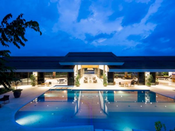 A luxurious poolside view of a modern building under a twilight sky, reflecting lights and surrounded by outdoor seating and greenery.