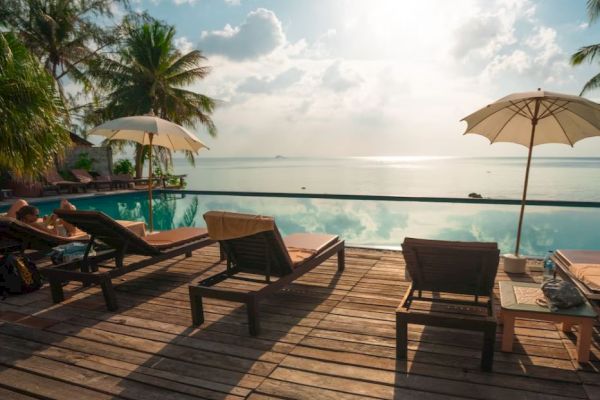 A serene poolside setting with lounge chairs, umbrellas, palm trees, and a view of the ocean under a bright sky at sunset or sunrise.