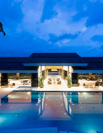 A luxurious pool area is leading to an inviting building under a twilight sky, with trees silhouetted in the foreground.