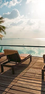 A tranquil poolside scene with lounge chairs, umbrellas, palm trees, and a view of the ocean under a partly cloudy sky.