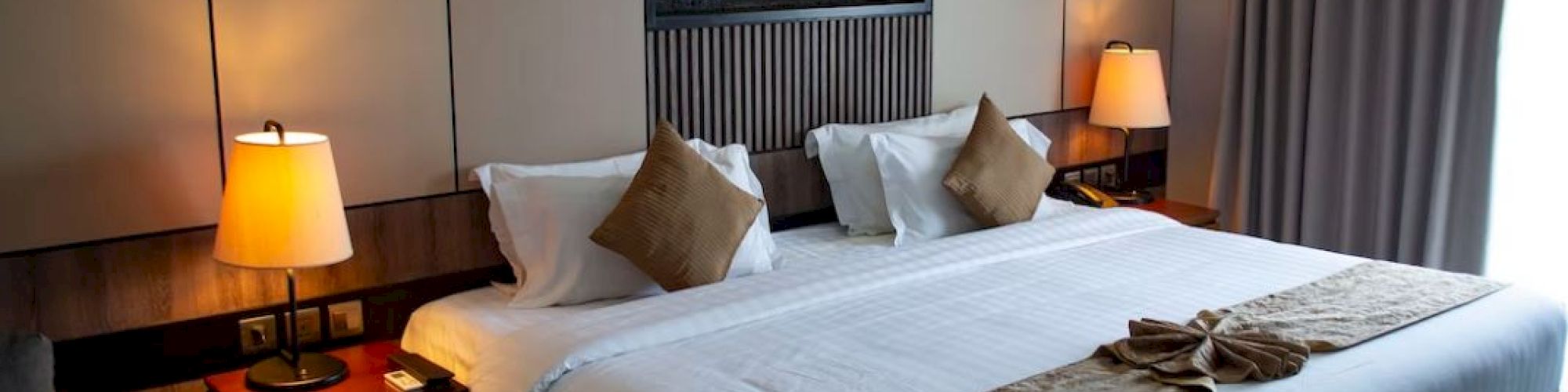 A neatly made hotel room bed with pillows, lamps on side tables, and a decorative fireplace feature on the wall above.