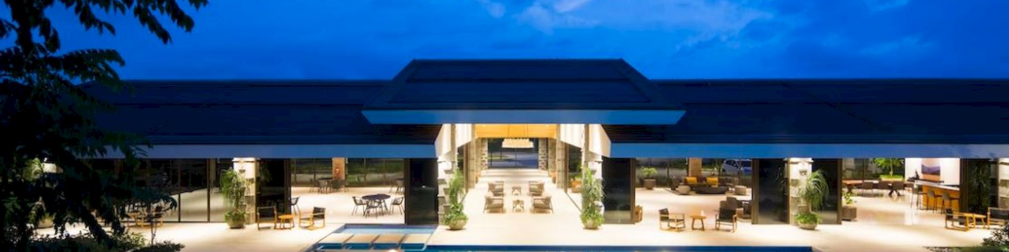 A luxurious building with a large illuminated outdoor pool, surrounded by seating and greenery, under a vibrant blue evening sky.