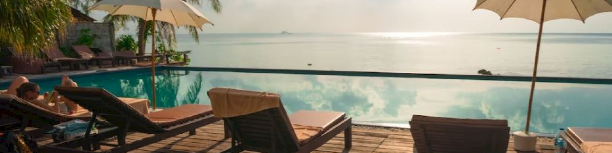 A serene poolside scene with lounge chairs, umbrellas, a wooden deck, and a backdrop of palm trees and a calm ocean under a bright sky.