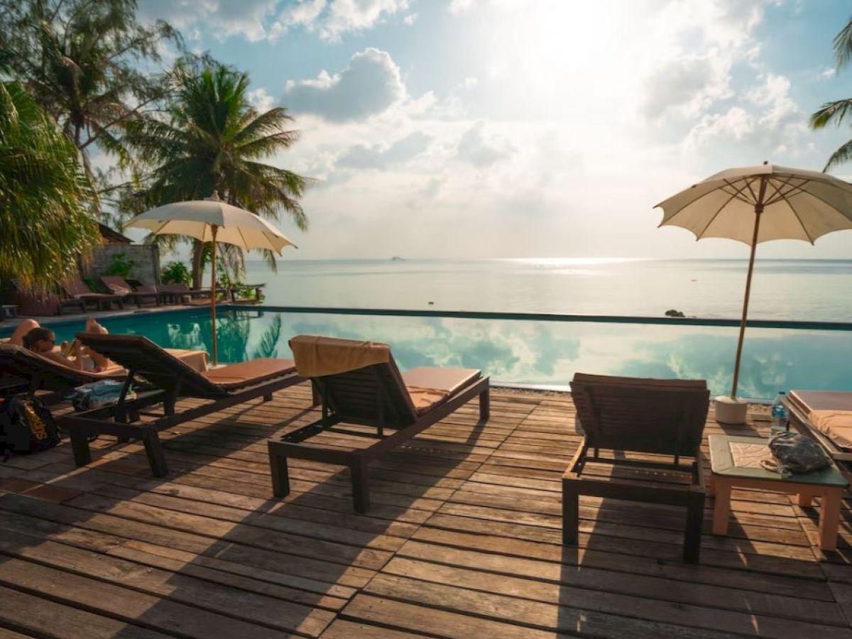 Poolside scene with lounge chairs, umbrellas, and palm trees overlooking a calm ocean and a cloudy sky at sunset.