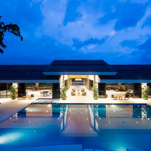 A modern building with a large, illuminated pool in the foreground, surrounded by seating areas under a deep blue evening sky.