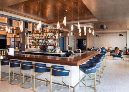 A modern bar with a well-stocked shelf, blue cushioned stools, hanging lights, and a relaxed seating area in the background.
