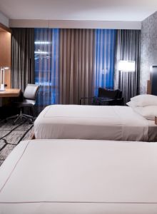 The image shows a modern hotel room with two beds, a TV, a work desk, and stylish decor.