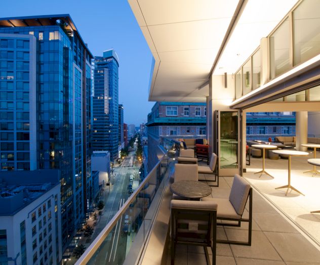 A modern balcony overlooks a cityscape at dusk, featuring tall buildings, seating areas, and warm lighting, creating an inviting atmosphere.