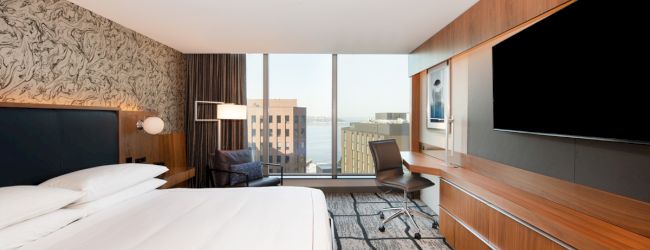 The image shows a modern hotel room with a large bed, desk, chair, TV, and city view through a window, featuring contemporary decor.