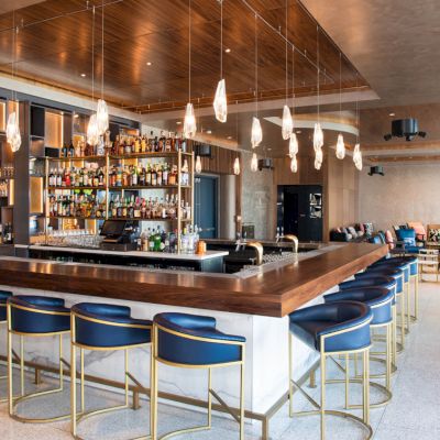A modern bar with blue chairs, stylish lighting, and a well-stocked shelf in a spacious, elegantly designed room.