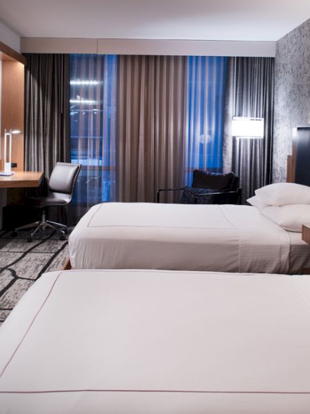 A modern hotel room features two beds, a desk with a chair, lamps, a wall-mounted TV, and a patterned accent wall, creating a cozy atmosphere.