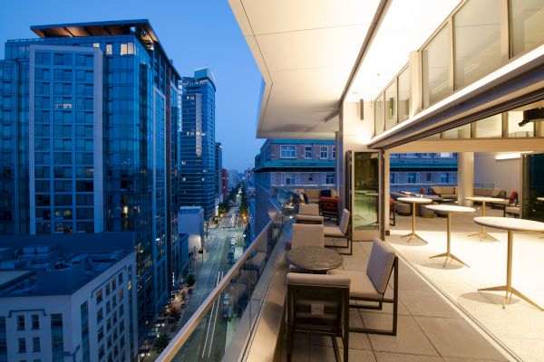 A modern cityscape at dusk, featuring a high-rise building with a furnished balcony overlooking a lit street with tall buildings.