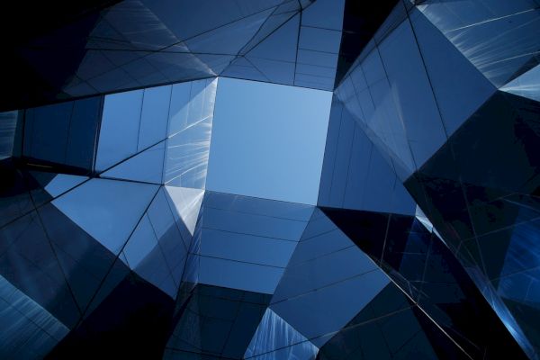 The image shows a geometric pattern of glass and metal, forming a kaleidoscopic view under an opening to the blue sky.