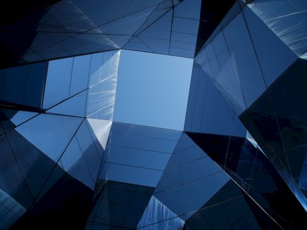 The image shows a geometric pattern of glass and metal, forming a kaleidoscopic view under an opening to the blue sky.