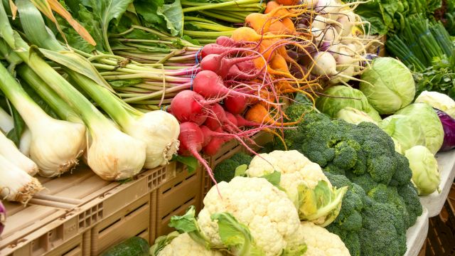 A colorful display of fresh vegetables: onions, radishes, carrots, broccoli, cauliflower, cabbage, and greens neatly arranged for sale.