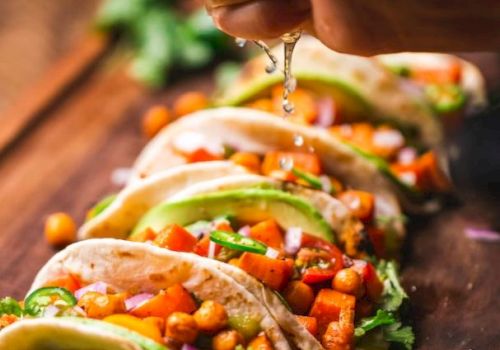A hand squeezes lime over a row of soft tacos filled with chickpeas, veggies, and garnished with cilantro and lime slices on a wooden board.