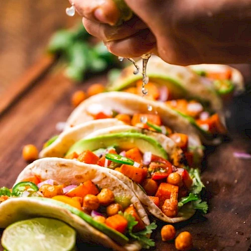 A hand squeezes lime over a row of soft tacos filled with chickpeas, veggies, and garnished with cilantro and lime slices on a wooden board.