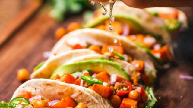 A hand squeezes lime over a row of soft tacos filled with chickpeas, veggies, and garnished with cilantro and lime slices on a wooden board.