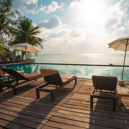 A tranquil beach scene with lounge chairs, umbrellas, and a pool overlooking the ocean under a bright sky with scattered clouds.