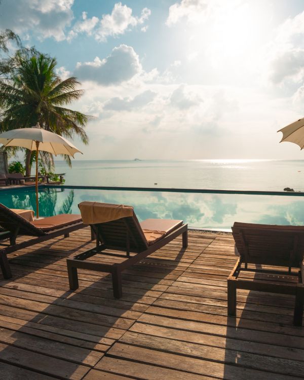 A tranquil beach scene with lounge chairs, umbrellas, and a pool overlooking the ocean under a bright sky with scattered clouds.