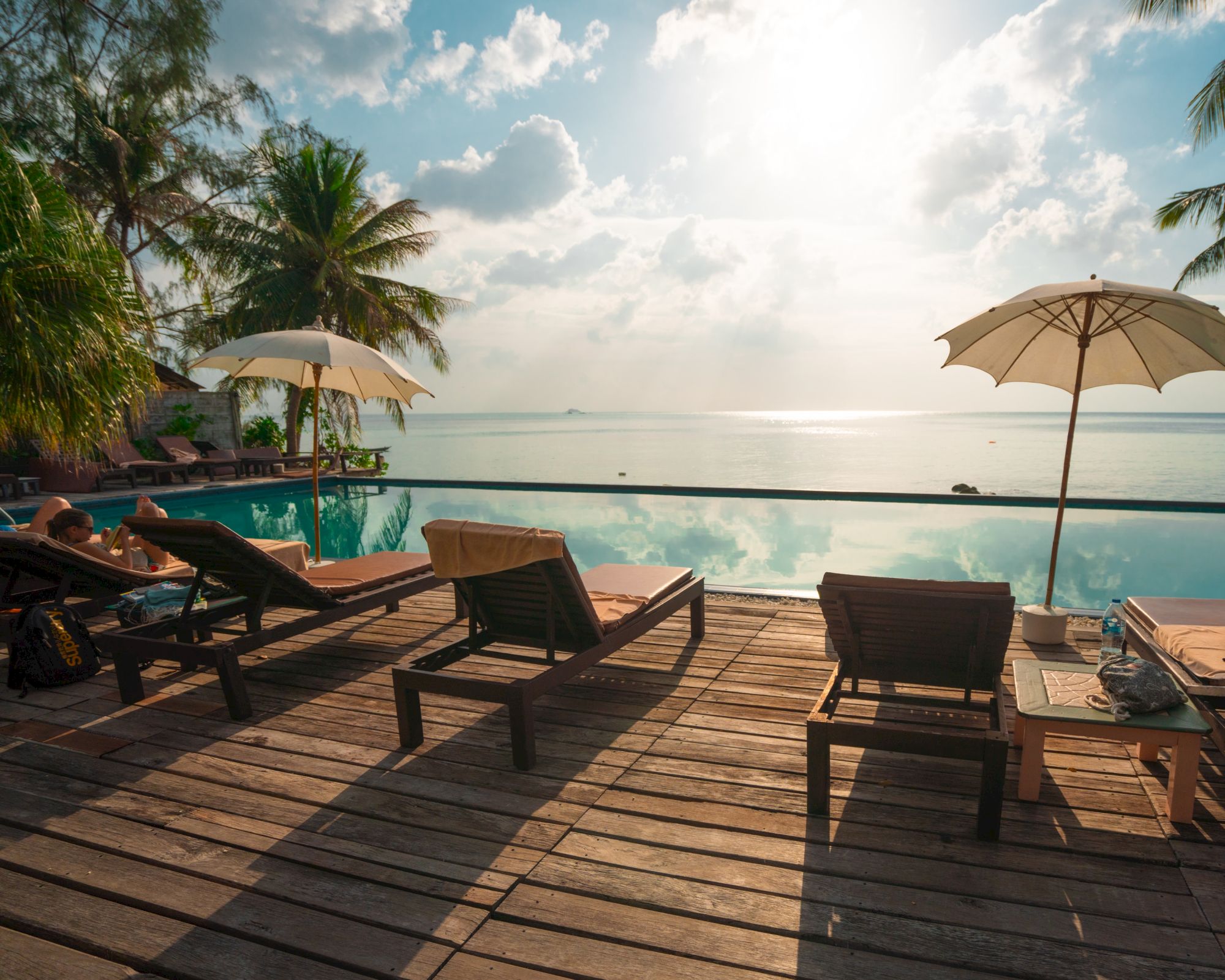 A serene beach scene with lounge chairs, umbrellas, and a pool overlooking the ocean, under a partly cloudy sky.