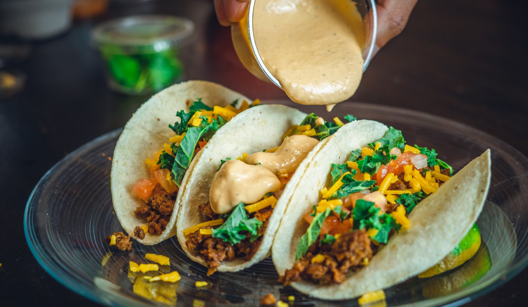Three tacos on a plate filled with meat, cheese, greens, and tomatoes, being topped with a creamy sauce.