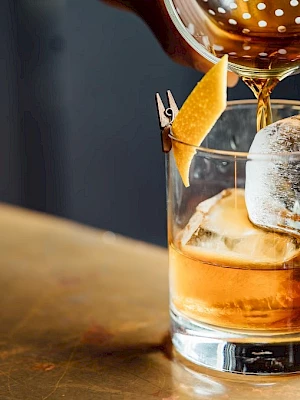 A cocktail being poured into a glass with a large ice cube and a twist of citrus peel on the rim.