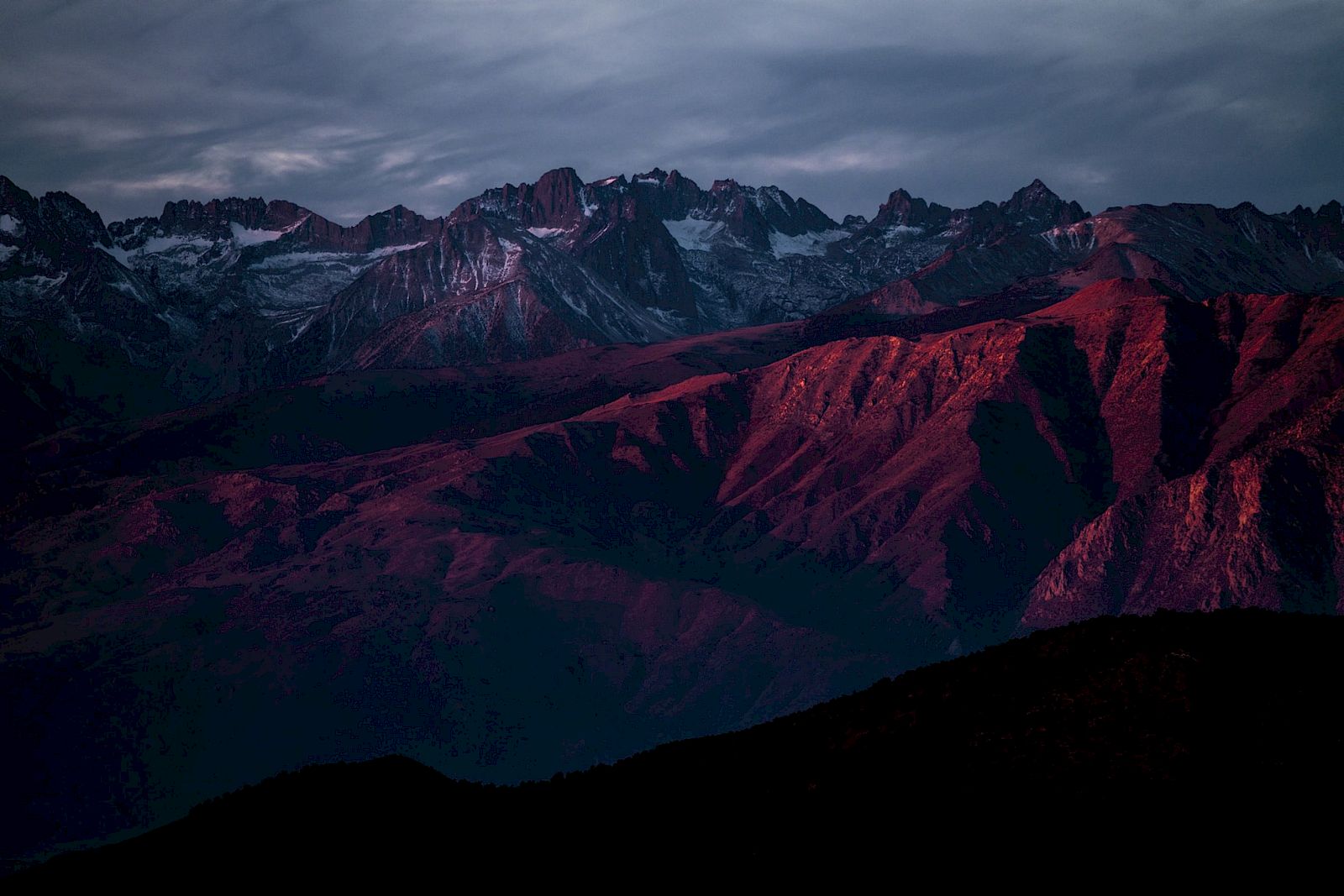 A mountain range at sunset with snow-capped peaks, bathed in a dramatic red and purple light, under a cloudy sky.