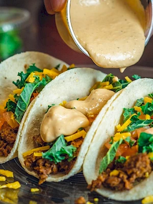Three tacos filled with ground meat, shredded cheese, lettuce, and salsa, being topped with a creamy sauce on a glass plate.