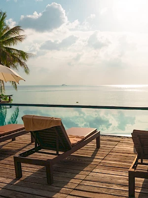 Lounge chairs, umbrellas, and a pool overlook a serene sea with palm trees under a bright sky.
