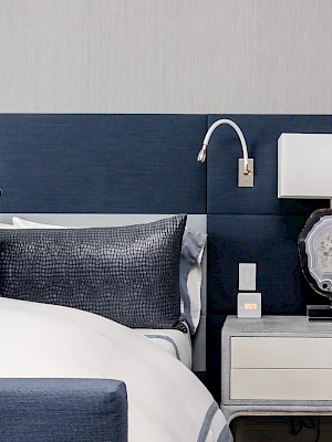 A modern bedroom features a bed with navy pillows and a nightstand with a lamp and book, set against a textured gray wall.