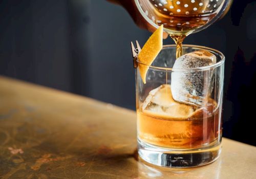 A hand pours a cocktail into a glass with ice and a citrus peel garnish, placed on a bar surface.