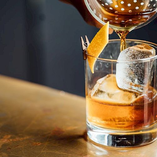 A hand pours a cocktail into a glass with ice and a citrus peel garnish, placed on a bar surface.