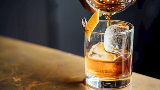 A hand pours a cocktail into a glass with ice and a citrus peel garnish, placed on a bar surface.