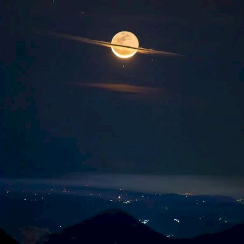 The image shows a full moon with clouds resembling Saturn's rings, above a landscape lit by distant city lights, creating a mystical scene.