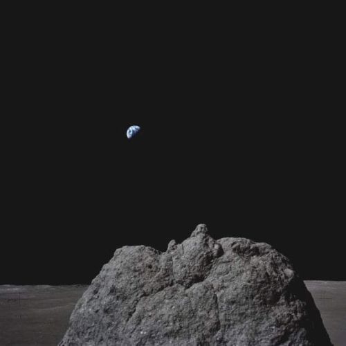 A rocky surface is seen with the Earth visible in the distant background against a dark sky.