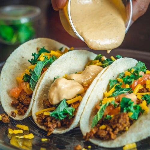 Tacos with ground meat, lettuce, tomato, and cheese, topped with a creamy sauce being poured over them.