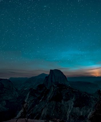 A starry night sky over a mountainous landscape, with the silhouette of a peak and a glowing horizon.