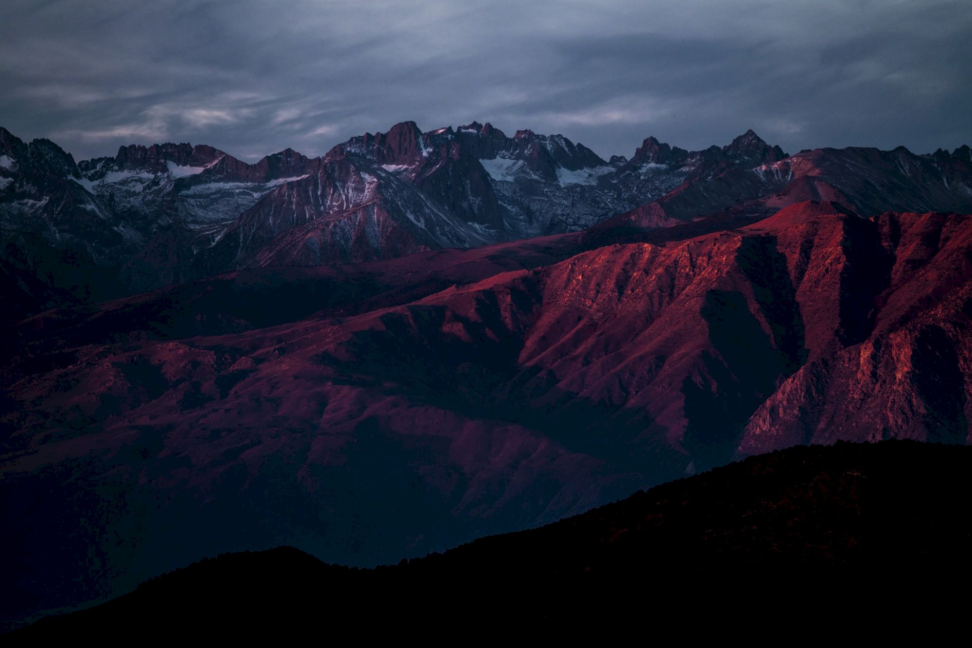 A mountain range at sunset with dark red hues on the slopes and dramatic, cloudy skies overhead.