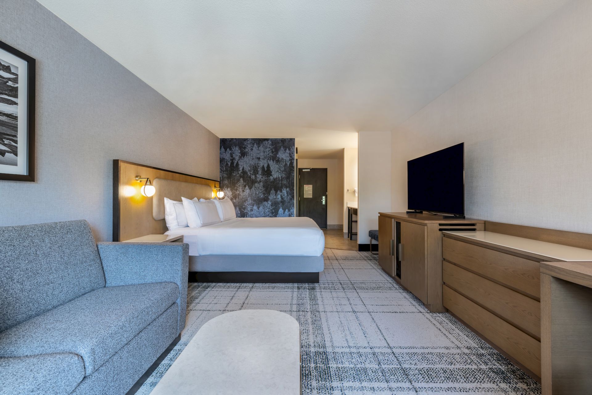 A modern hotel room with a king bed, sofa seating, a TV, and forest-view window, creating a calm, cozy stay.
