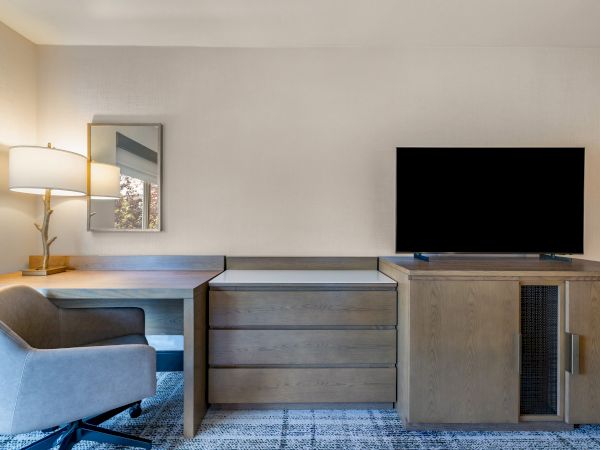 A modern room with a desk, lamp, chair, mirror, and TV on a wooden cabinet, all set against a light wall backdrop.