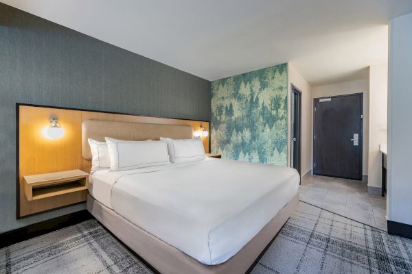 A modern hotel room with a large bed, stylish headboard, tree-patterned accent wall, and a door in the corner.