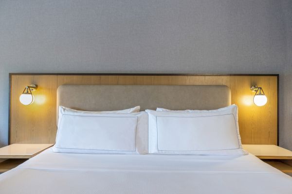 A neatly made bed with white pillows and sheets, two bedside lamps are mounted on a wooden headboard.