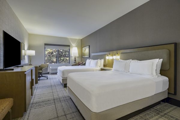 The image depicts a hotel room with two large beds, a TV, a desk with a chair, and lamps, all in a modern and spacious setting.