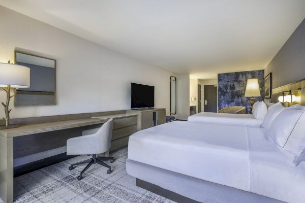 A hotel room with two beds, a desk with a chair, a TV, lamps, and a patterned wall near the door and mirror.