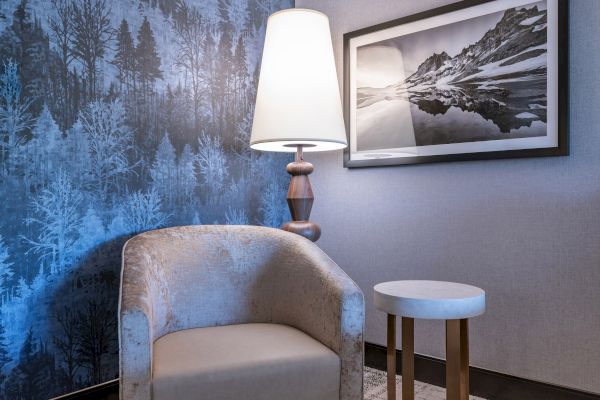 Room with a cozy chair, tall lamp, framed mountain photo, and blue forest-themed wallpaper.