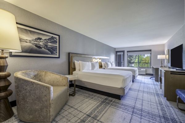 A modern hotel room with two beds, a sitting area, desk, TV, and large window overlooking greenery.
