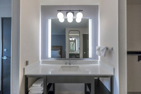 A modern bathroom vanity with a sink, mirror, towel, and overhead lights, featuring a reflection showing another room with a bed.