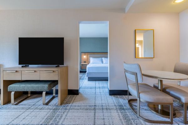 The image shows a hotel room with a TV, desk, chair, and a mirror, leading to a bedroom with a bed and lamp through an open doorway.